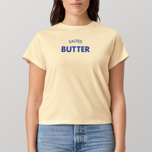 A woman wearing a pale yellow short-sleeve t-shirt with 'SALTED BUTTER' printed in blue text on the chest, paired with blue jeans, against a solid gray background.
