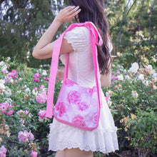 A woman in a white dress stands in a garden, viewed from behind, carrying a pink floral shoulder bag with a bright pink strap.