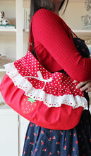 A red shoulder bag with white polka dot fabric, white lace ruffles, and a strawberry applique, carried by a person wearing a red ribbed sweater and a navy blue strawberry-print skirt.