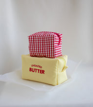Two fabric cosmetic pouches stacked on a white surface against a white draped backdrop. The bottom pouch is pale yellow with red text reading 'UNSALTED BUTTER,' and the top pouch is red-and-white gingham with a red zipper pull.