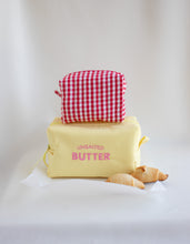 A yellow canvas lunch bag with pink text reading 'UNSAUTED BUTTER' sits on a white surface with a red-and-white gingham pouch stacked on top. Two croissants are placed in front, against a soft white draped background.