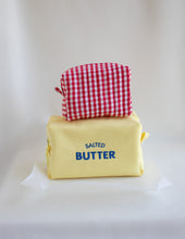 A novelty yellow bag styled like a butter package with blue 'SALTED BUTTER' text, topped with a red-and-white gingham pouch, set against a draped white fabric background.