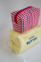 Two zippered cosmetic pouches stacked on a white surface. The top pouch is red and white gingham with a pink zipper, and the bottom pouch is pale yellow with blue text reading 'SALED BUTTER'.